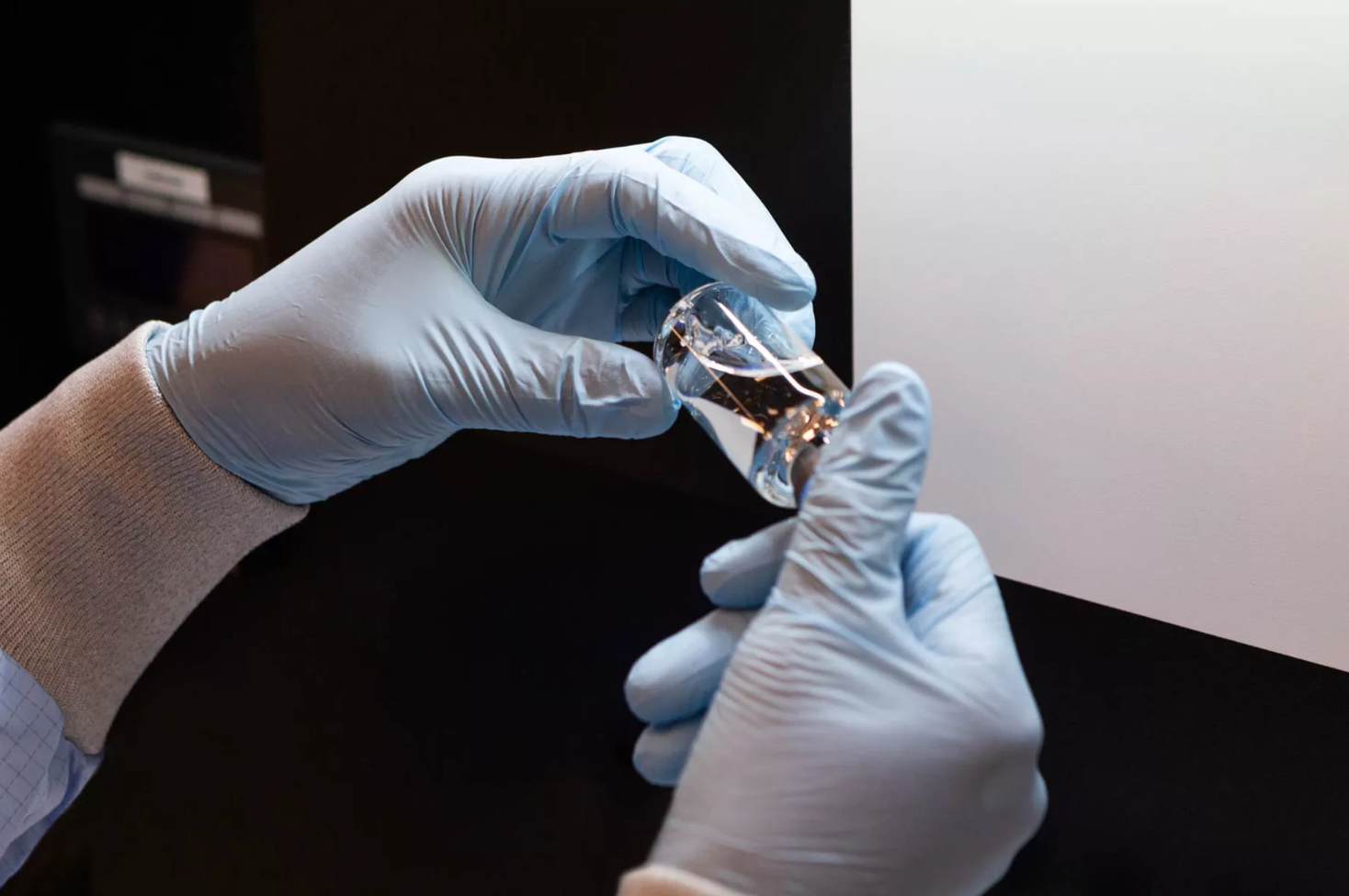 A close up of a medical professional's hands wearing blue surgical gloves holds a small glass bottle of a new anti-viral drug in a lab setting. 